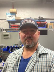 Roger Nadeau with machinery and equipment in the background, wearing a hat with "Veteran" on it.