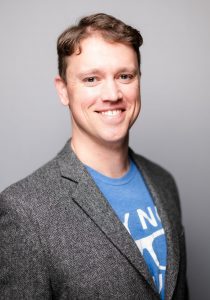 Jacob Johnson smiling, wearing a blue t-shirt under a blazer.