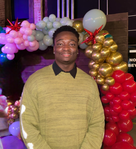 Justin Tomlinson standing in front of a stand of multi-colored balloons wearing a light green sweater with black collared shirt underneath.