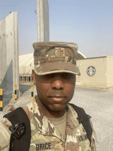 Ulrick Brice dressed in fatigues standing on base overseas in front of a building with a Starbucks logo on a clear sunny day.