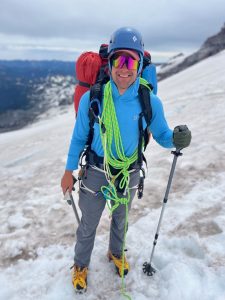 Alex Sayre standing on a snow covered mountainside in winter gear with views of mountains in the background.