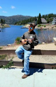 Eddie Langarica leaning on a fence in front of a river, playing a ukelele on a sunny day.